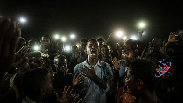 مردم در حال خواندن شعار هستند، در حالی که یک جوان شعری را که توسط تلفن های همراه روشن می شود ، در اعتراضات در خارطوم سودان 2019 می خواند. این عکس توسط یاسویوشی چیبا برای آژانس خبری فرانسه گرفته شده است.